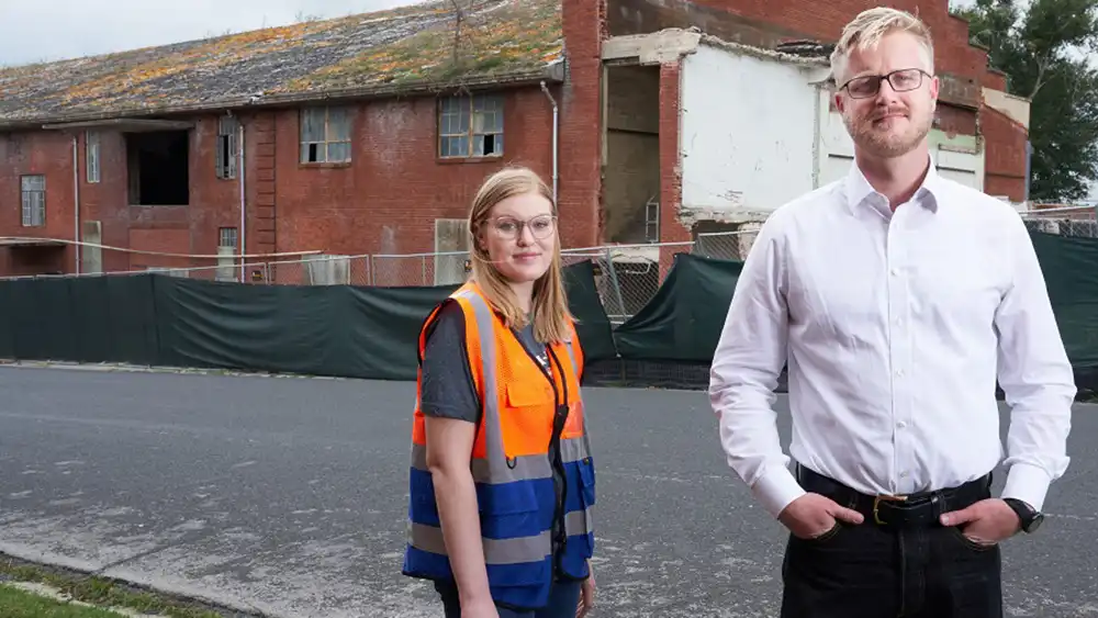 A man standing with his hands in his pockets, next to a woman in a construction vest.