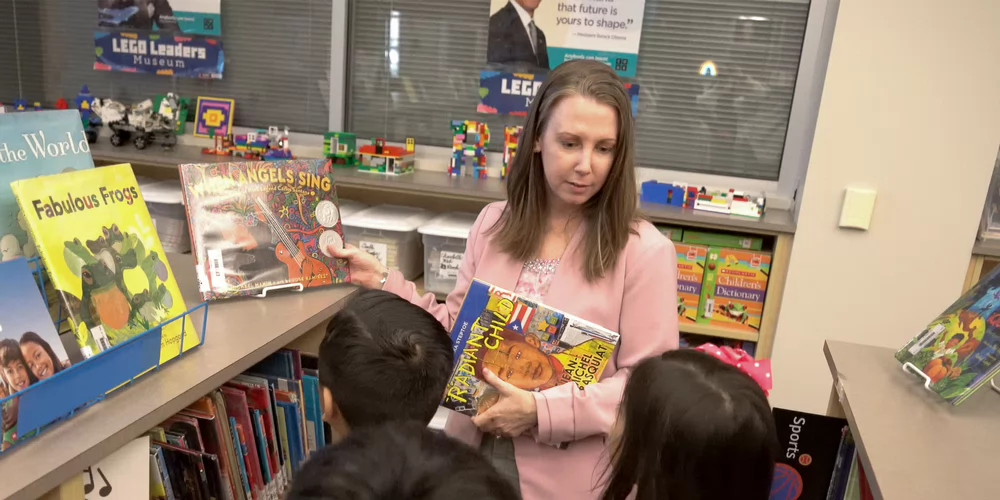 Sam Houston State University (SHSU) library science and technology program, shows a librarian holding children's books while interacting with young students in a library.
