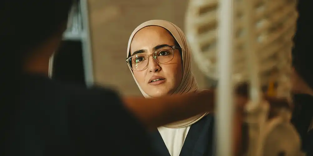 SHSU College of Osteopathic Medicine student doctor in a hijab and glasses attentively studies a human skeleton model during a hands-on lab session