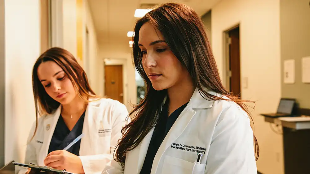Two student doctors one standing behind the other one with a clipboard taking notes