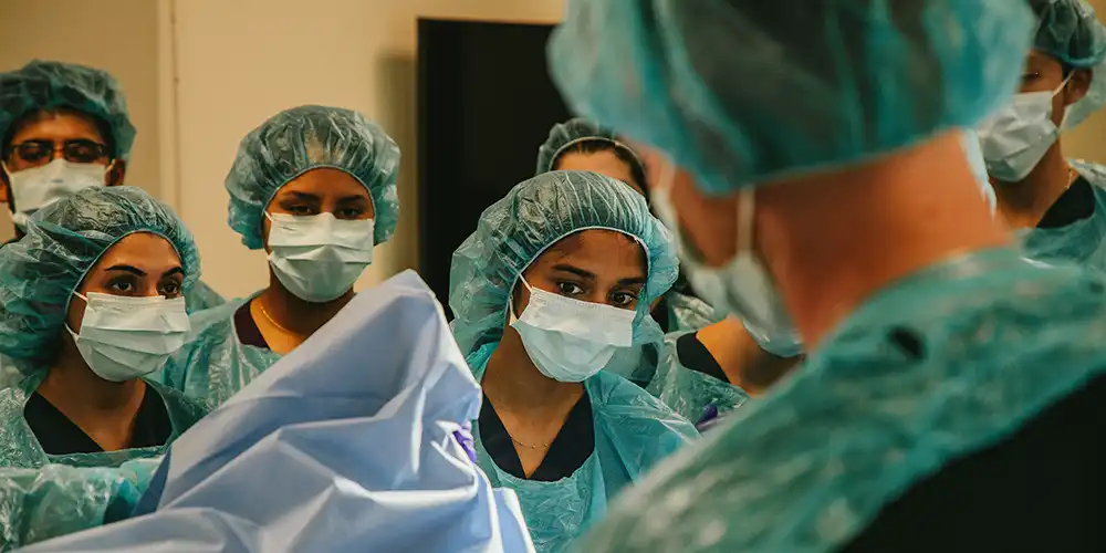 A group of medical students in scrubs, masks and surgical caps watching a medical demonstration in a simulation hospital room