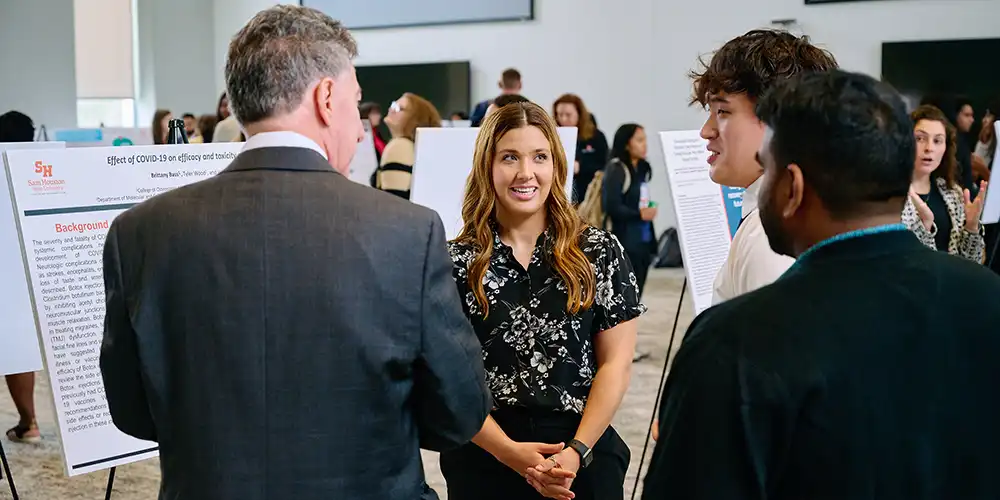 Student facing camera and presenting research poster to a group of three people with backs to camera