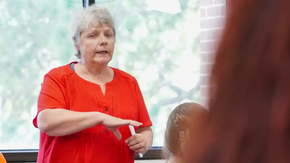 Professor speaking to the camera in a classroom style class, with her hands opened.