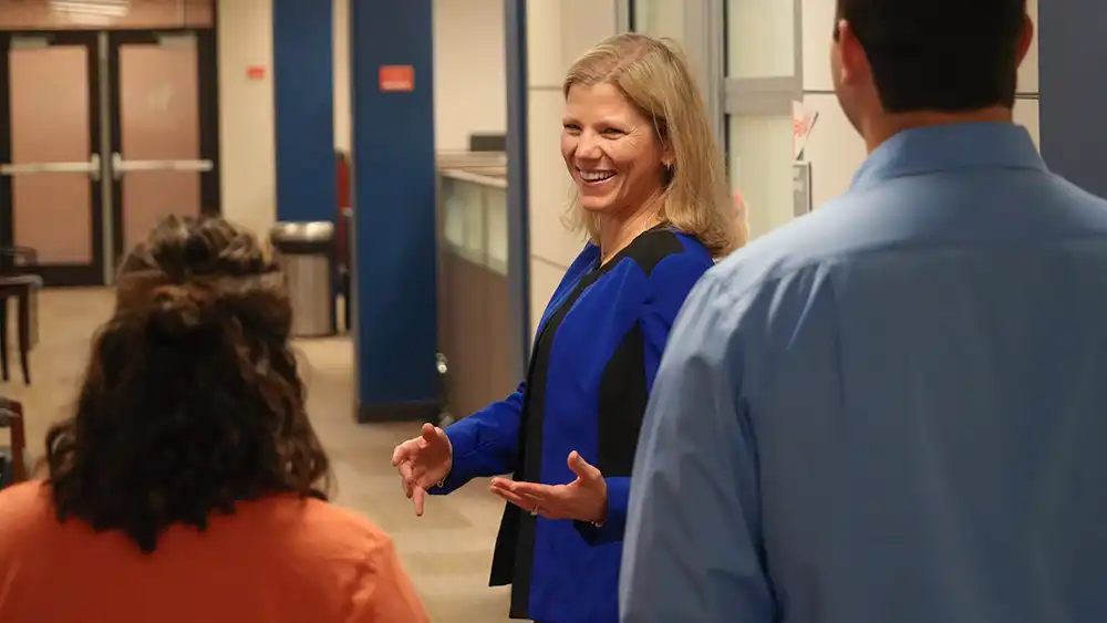 Lady in royal blue suit walking down a hallway looking back over her shoulder smiling talking to two people