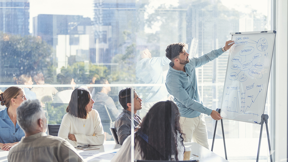 Man instructing at a whiteboard while group is listening.