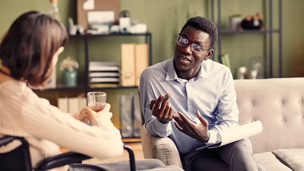 At a counseling session, a therapist is speaking to his client with a notebook on his lap and a pen in his hand, while his client in in a wheelchair holding a cup of water.