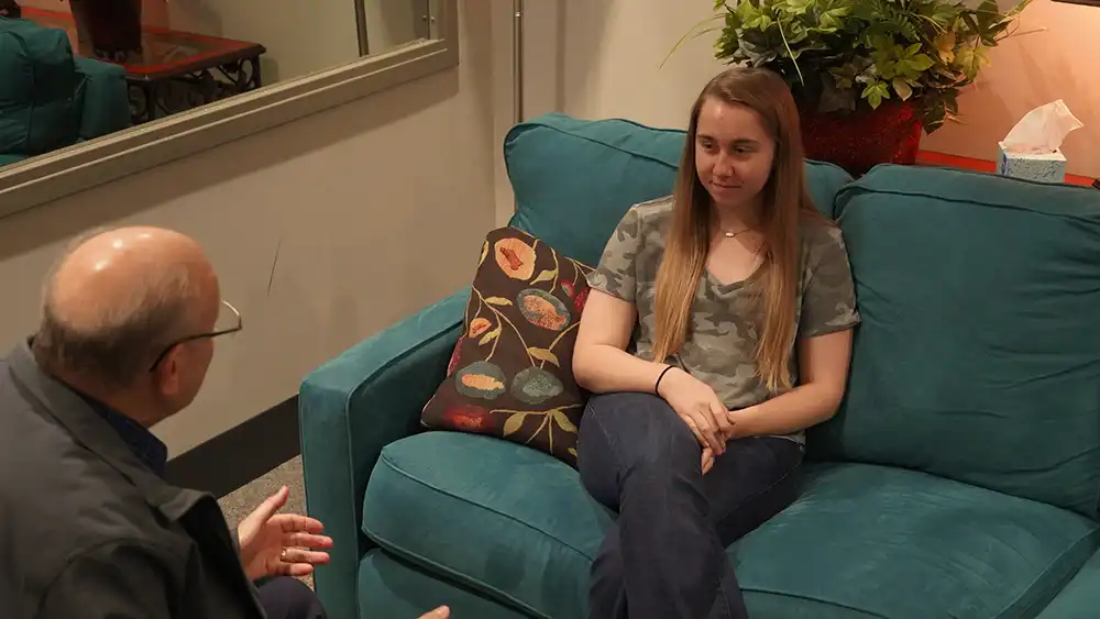 At a counseling session, a therapist is talking to his client, while she sits and smiles on a couch