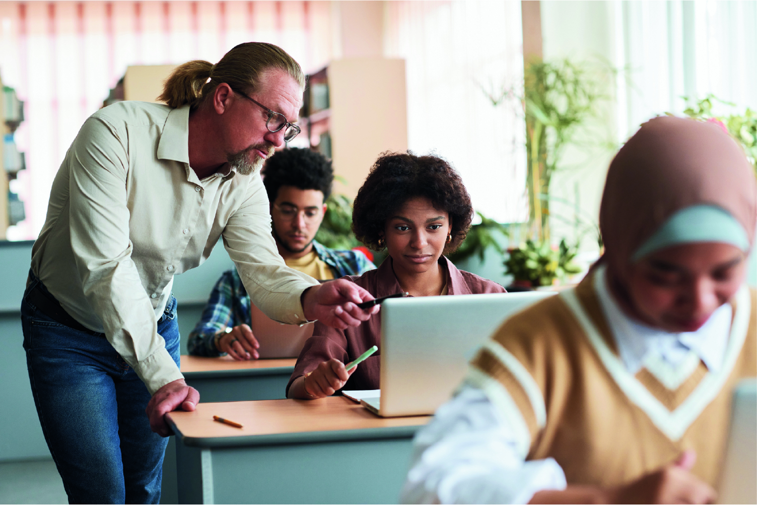 Man in a classroom leaning on student’s desk pointing to her computer screen .