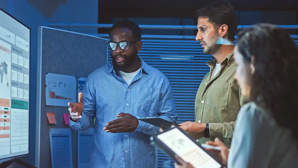Three people in a modern office setting are gathered around a large monitor displaying charts, graphs, and data visualizations. One person is gesturing toward the screen while another holds a tablet, and a third person is partially visible in the foreground. The background includes a bulletin board with papers and sticky notes, and the lighting is dim, creating a focused work environment.