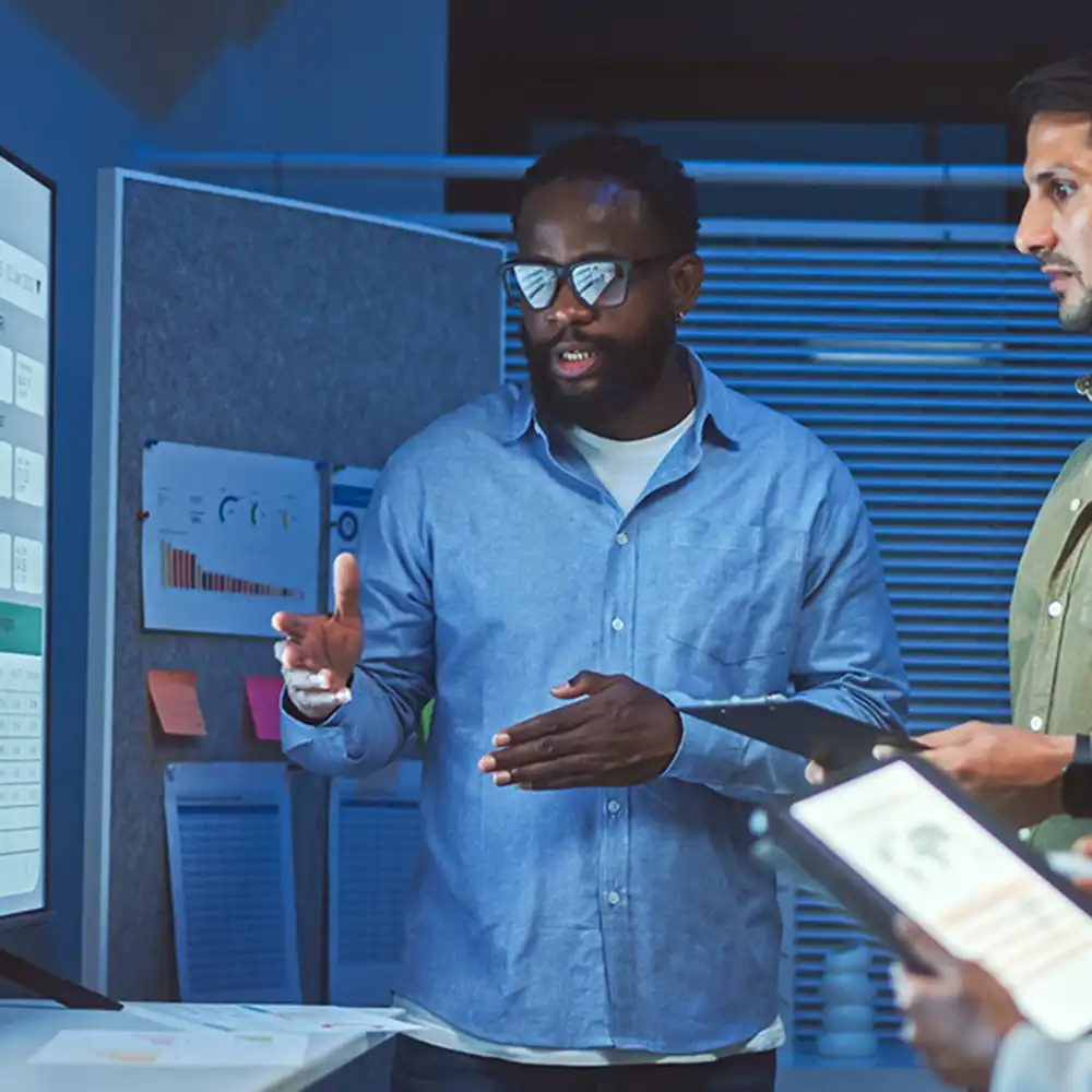 Two people standing in front of a large monitor displaying charts and data, engaged in a discussion. One person is gesturing toward the screen while the other holds a tablet. The background includes a bulletin board with papers and sticky notes, and the setting appears to be a modern office or meeting space with dim lighting.