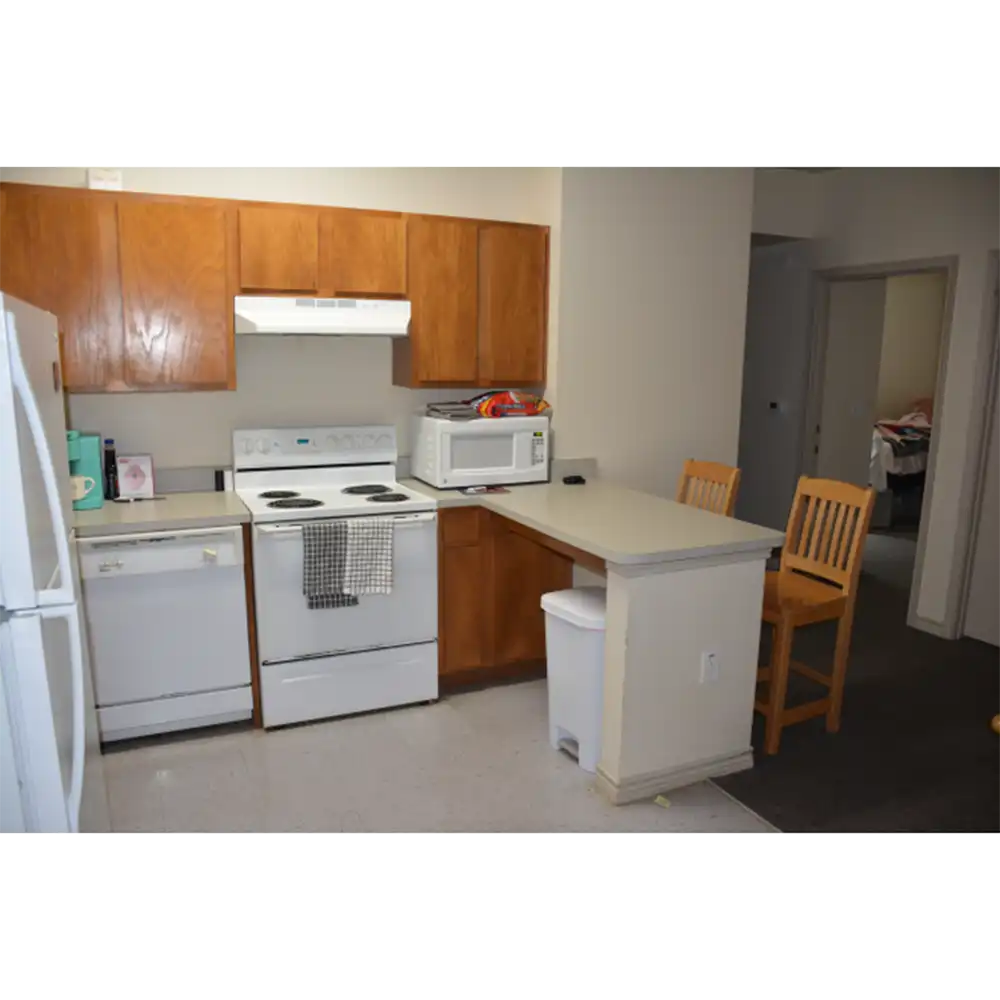 A kitchen in SHSU's Bearkat Village Apartments featuring wooden cabinets, a white electric stove with an oven, a white range hood, a microwave on the countertop, and a dishwasher. There is also a refrigerator on the left side and two wooden bar stools at the counter. The kitchen has beige walls and tile flooring.