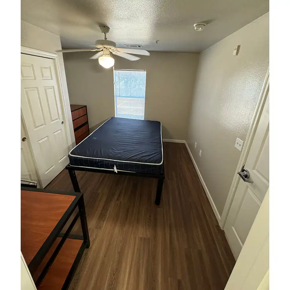 An image of the bedroom at SHSU's Campus Edge Apartments with beige walls and wooden flooring. A double bed with a dark blue mattress is positioned against the right wall beneath a ceiling fan with a light fixture. A window with closed blinds is centered on the far wall. To the left of the bed is a wooden dresser with three drawers and a closet with white sliding doors. A desk or table is partially visible in the foreground, and two electrical outlets with a light switch plate are on the right wall near the door.