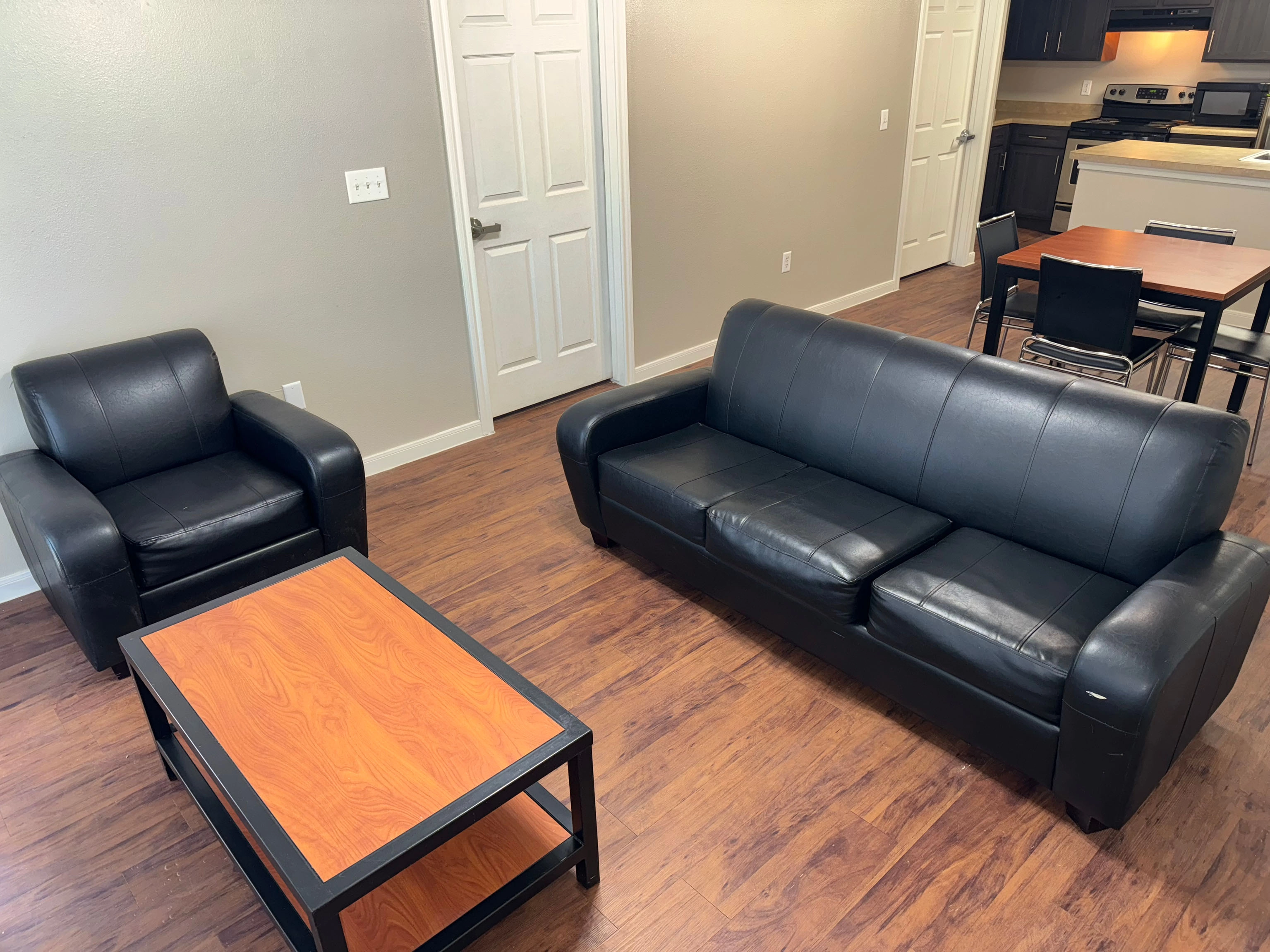 Living room at SHSU's Campus Edge Apartments featuring a black leather sofa and matching armchair on a wooden floor. A rectangular coffee table with a wooden top and black frame sits in front of the sofa. In the background, an open kitchen area includes dark cabinets, stainless steel appliances, and a small dining table with four chairs. Two white doors are visible on the far wall, and the walls are painted a light neutral color.