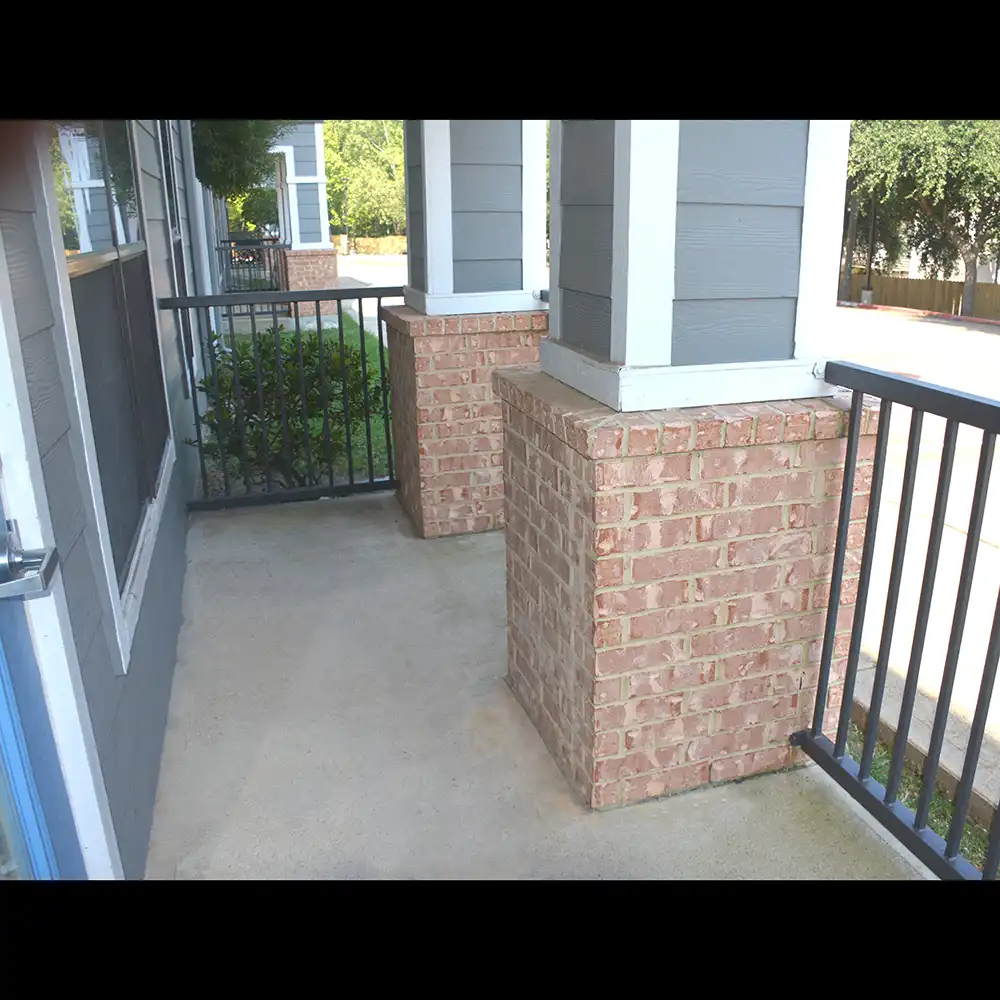 Outdoor patio area at SHSU's Campus Edge Apartments with concrete flooring, brick column bases, and black metal railings attached to a gray and white building exterior.