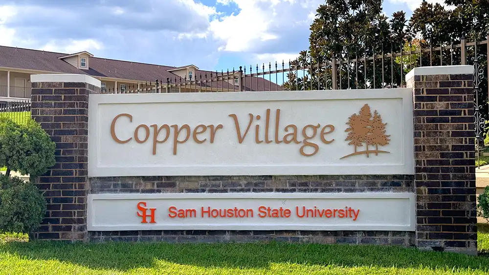 Exterior view of Copper Village Apartments at Sam Houston State University. A brick and stone sign in the foreground displays 'Copper Village' in large copper-colored letters on a white background, with an illustration of two trees to the right. Below, 'Sam Houston State University' is written in smaller red letters next to the university's orange 'SH' logo. In the background, a residential building with beige siding and a brown roof is partially visible behind a wrought iron fence and trees. The sky is partly cloudy.