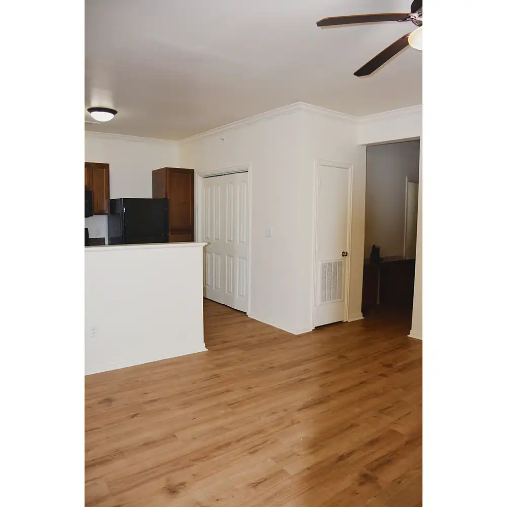 The living room of a 2-bedroom apartment at SHSU's Copper Village Apartments features light-colored walls and wood flooring. A ceiling fan with lights is mounted overhead. To the left, a partial view of the kitchen shows dark wooden cabinets and black appliances, including a refrigerator. Two white doors are visible—one appears to be a closet, and the other leads to another room or hallway.