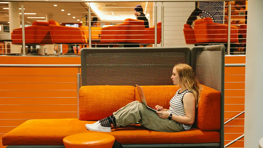 A student sits comfortably on an orange lounge sofa inside the Lowman Student Center (LSC), working on a laptop. The student is leaning back against a cushioned panel, with legs stretched out along the seat. In the background, other students sit and study on orange furniture on the upper level of the LSC.