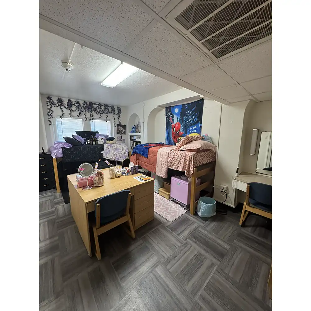 Bedroom in SHSU’s Belvin-Buchanan Hall with two lofted beds—one with black bedding and one with pink bedding. Each bed has storage underneath, and two central desks holds a mirror, makeup brushes, and books. A Spider-Man poster hangs above the pink bed. The room has gray herringbone tile flooring and two windows with floral curtains.
