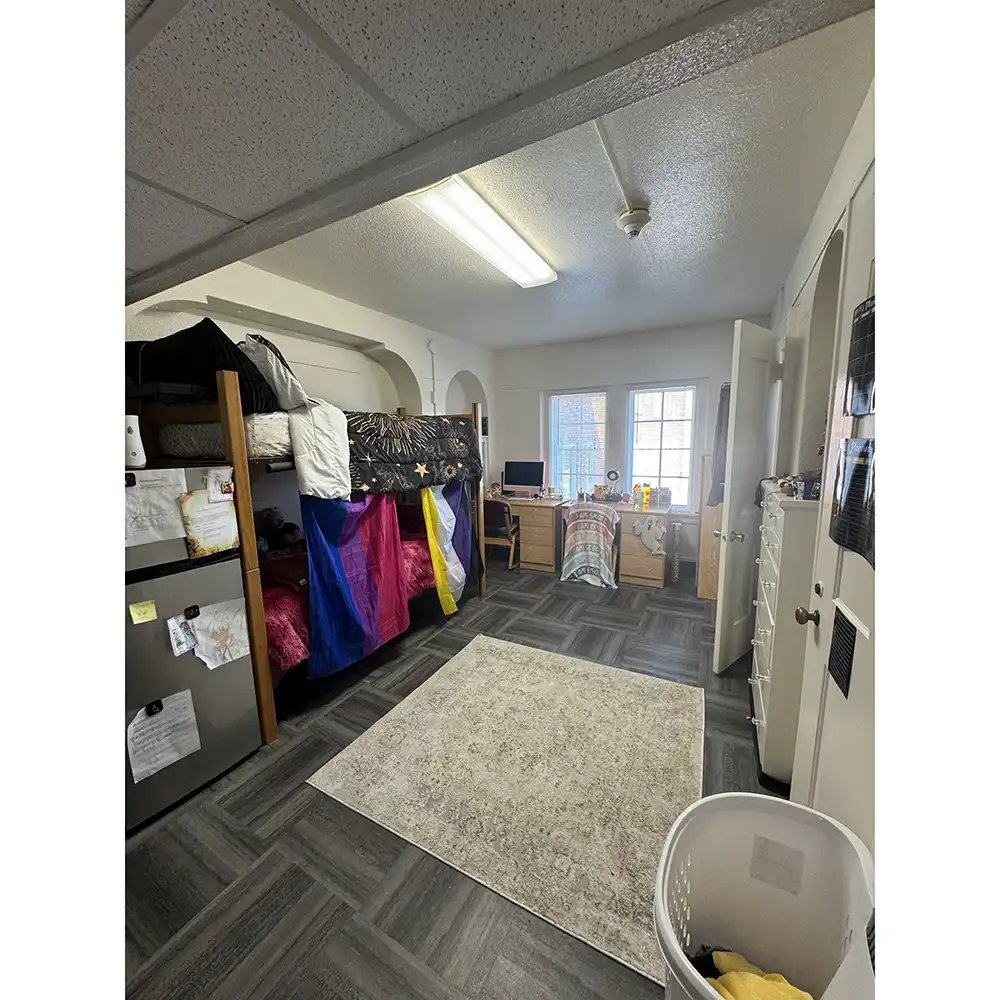 Bedroom in SHSU’s Belvin-Buchanan Hall with a lofted bed and colorful curtains creating a private space below. A desk with a computer sits beneath two large windows. The room has dark gray flooring, a small rug, storage units, and an open door leading to another area. Fluorescent lighting is mounted on a slightly slanted ceiling.
