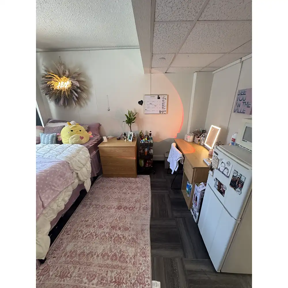 Bedroom in SHSU’s Jackson-Shaver Hall featuring a bed with pink bedding and plush toys, including a large yellow duck. A neon 'ALOHA' wreath hangs above the bed. A wooden nightstand with plants, a drink shelf, a desk with an illuminated mirror, and a mini-fridge with a microwave are also present. The room has dark tile flooring and a pink patterned rug.