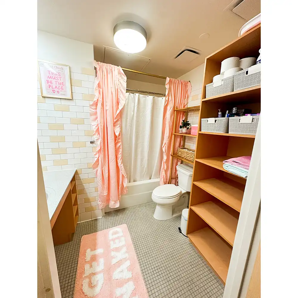 A residential bathroom in SHSU's Lone Star Hall featuring a shower with a white curtain and pink ruffled outer curtains, a toilet, and wooden shelving units. The shelves are stocked with various items including toilet paper, towels, and storage baskets. The floor is tiled in small gray squares, and there is a pink bath mat with the words 'GET NAKED' in white letters. The walls have light-colored tiles arranged in a staggered pattern. A framed sign above the counter reads 'This must be the place