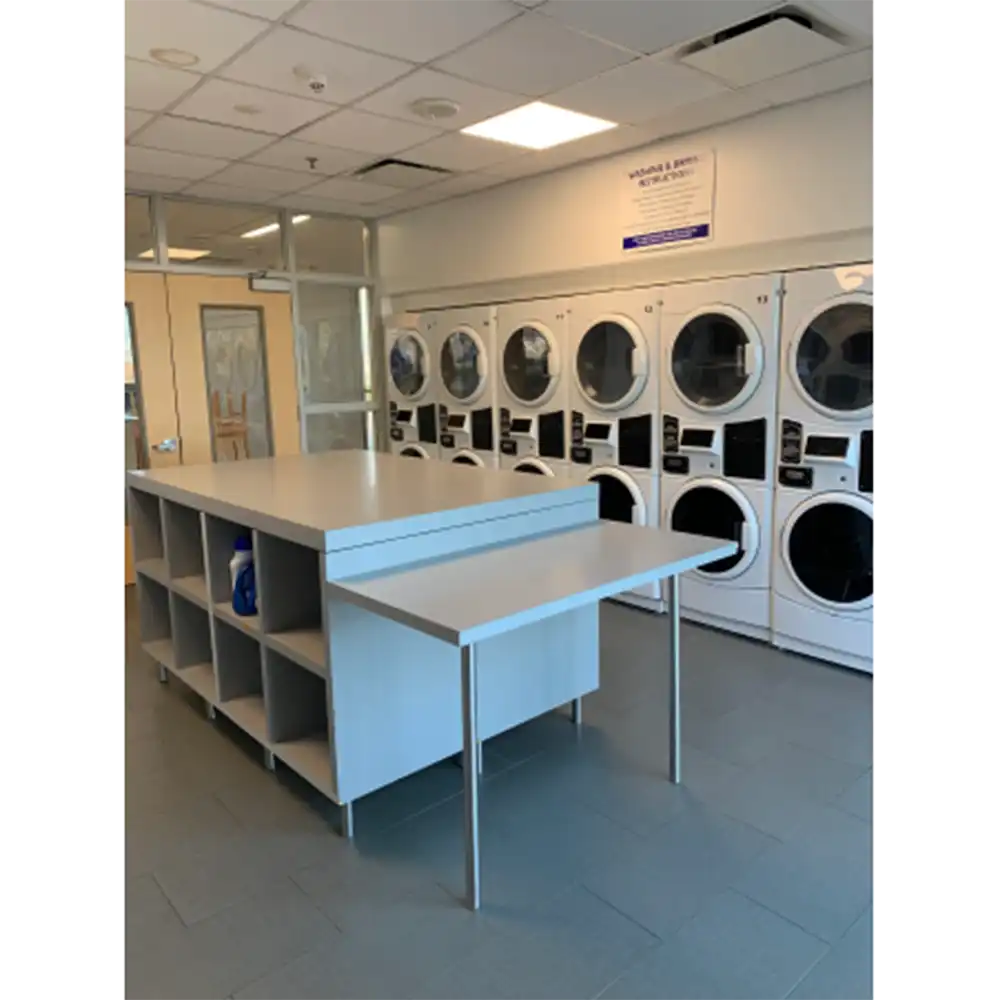 Laundry room in SHSU’s Lone Star Hall with a row of front-loading washers and dryers along the back wall, a large folding table with open shelving in the center, tiled flooring, recessed lighting, and an instructional sign on the wall.