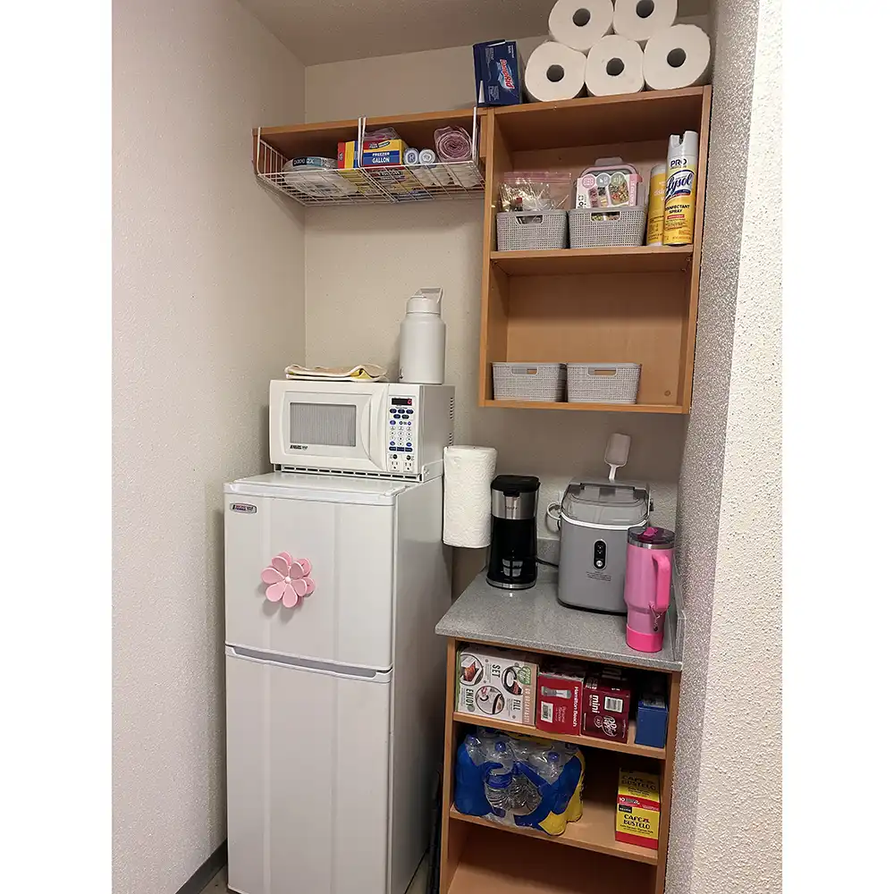 Pantry area in SHSU’s Lone Star Hall bedroom features a white refrigerator with a pink flower magnet, a microwave on top, and open shelving above with paper towels and kitchen supplies. A nearby countertop holds a coffee maker, ice machine, and tumbler, with additional shelves storing food items, cleaning supplies, and bottled water.