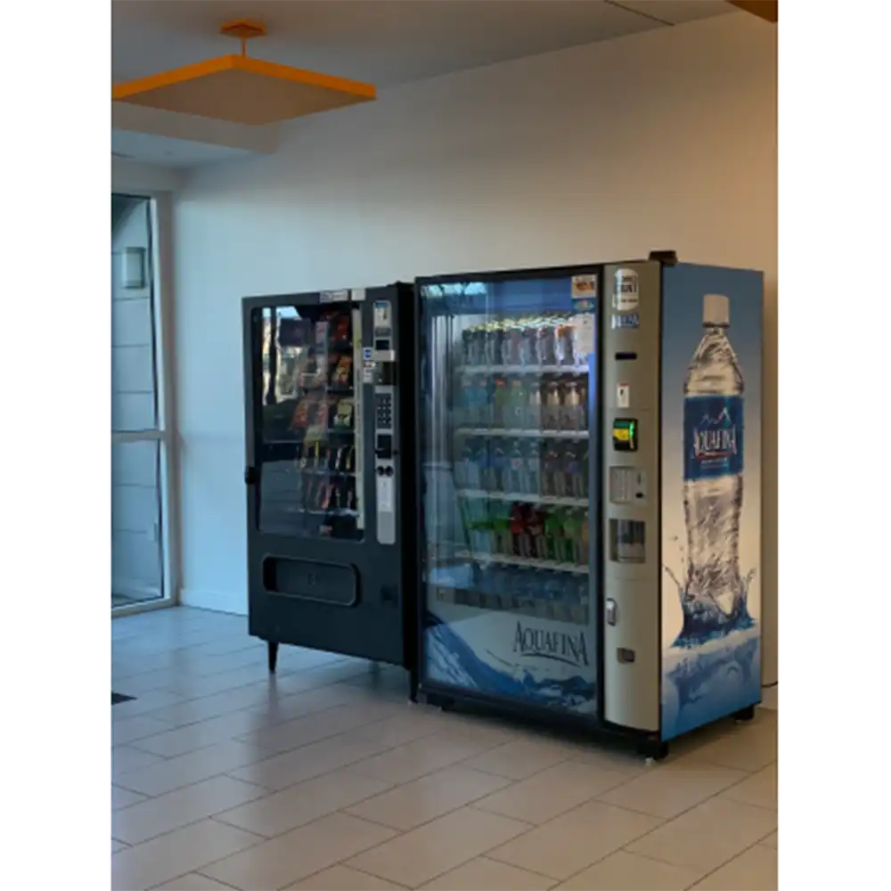 Two vending machines on the 1st floor of SHSU’s Lone Star Hall: a black snack machine with visible snack items and an Aquafina-branded drink machine with bottled beverages. Both are set against a white wall with tiled flooring.