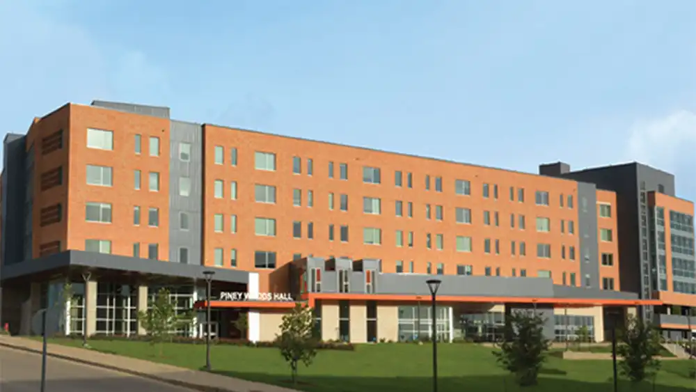 Large, modern five-story residence hall named "Piney Woods Hall," featuring red brick and dark gray accent panels. The building has a glass-walled entrance with a covered walkway and is surrounded by a landscaped lawn with young trees and streetlamps along a gently curving road.