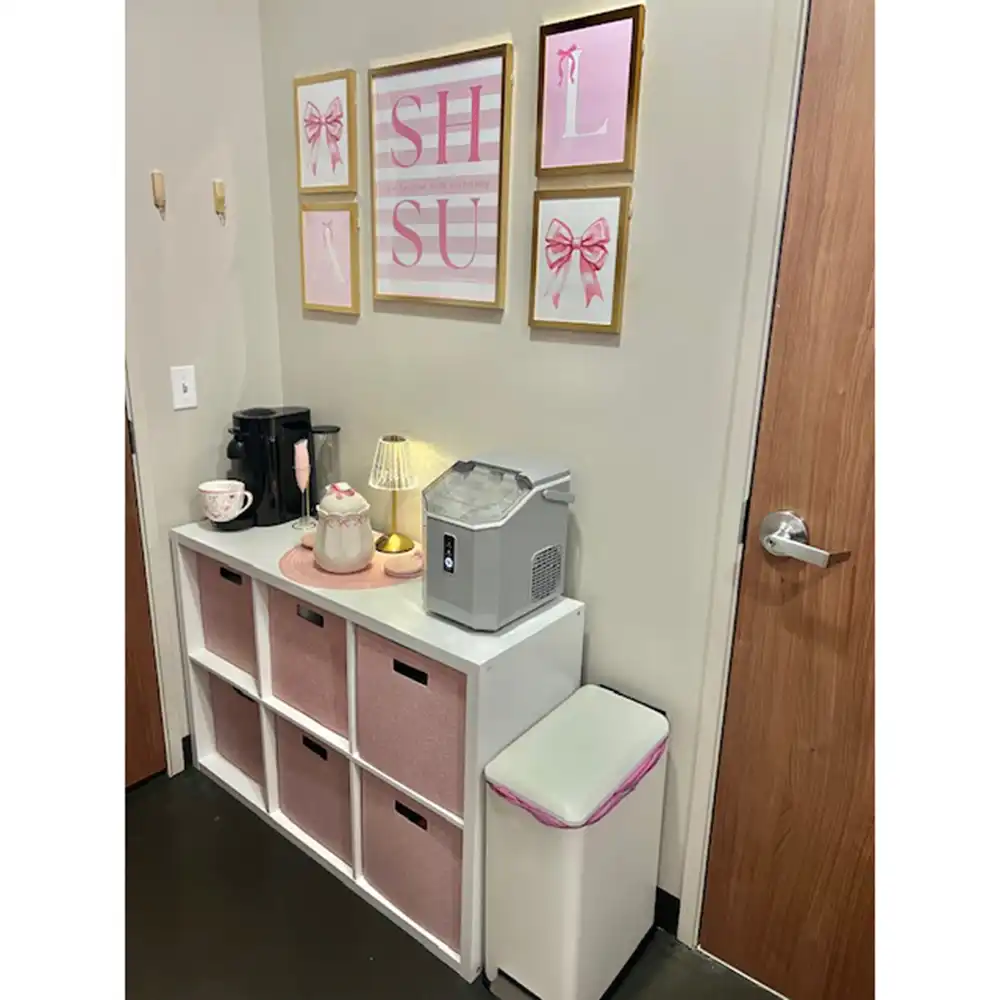 Entrance to one of the rooms at Piney Woods Hall featuring a white six-compartment storage unit with pink fabric bins. On top are a gold-based lamp, an ice maker, a teapot with cups, and decorative items. Above the unit are five framed pictures with pink and white designs, including one that says 'SHSU.' A white trash can with a pink-rimmed lid is to the right, next to a wooden door. The walls are light-colored.