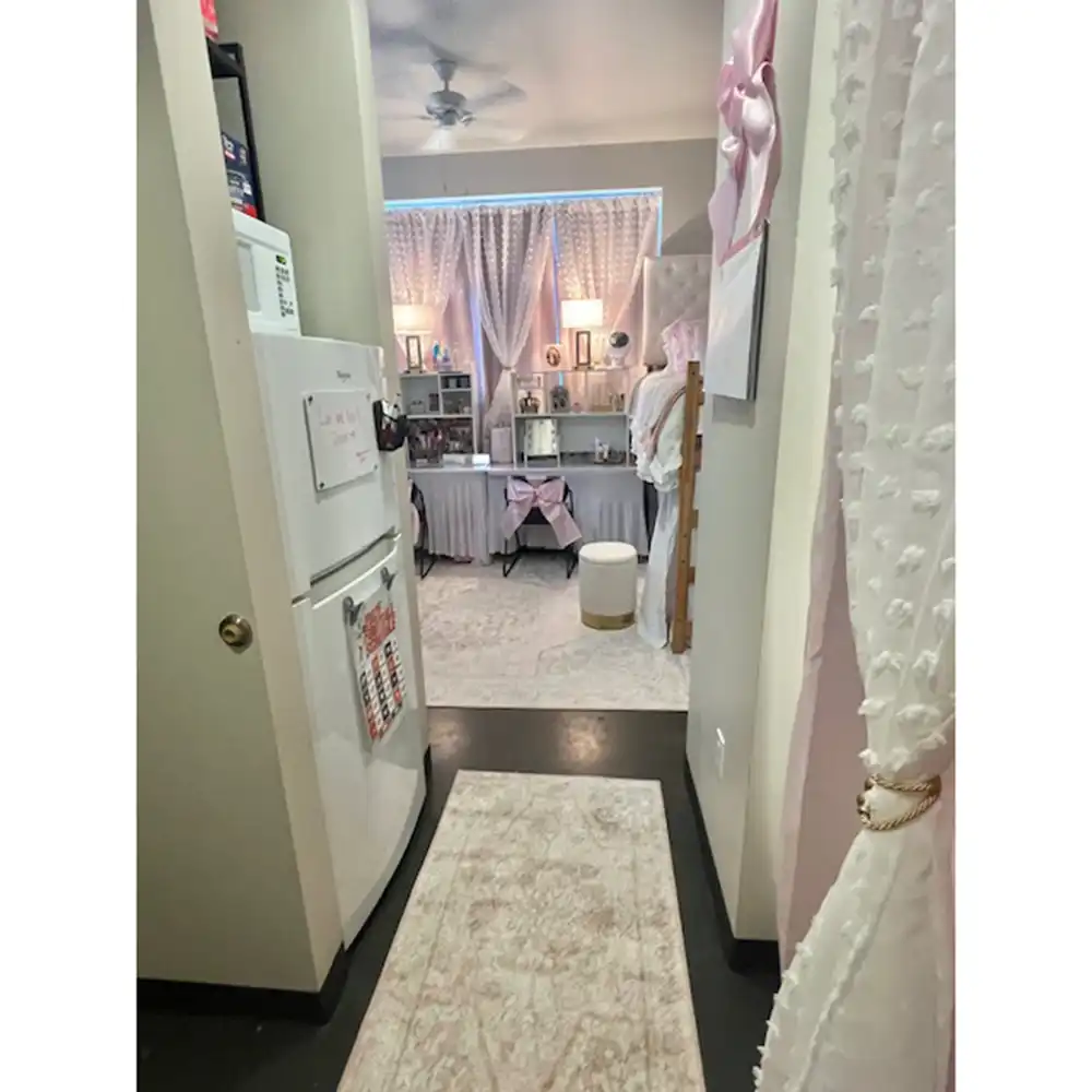 Microwave and refrigerator area in SHSU's Piney Woods Hall featuring a white Whirlpool refrigerator with a calendar and small whiteboard on the front. A white microwave sits above the refrigerator with items stored on top. The area opens into a dorm room decorated in pink and white, with curtains, lamps, and furniture visible.