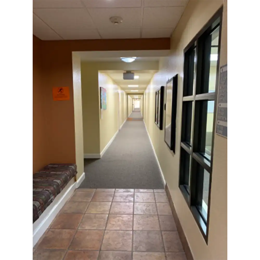 Interior hallway of SHSU's Raven Village featuring beige walls, a light-colored ceiling, and flooring that transitions from tile to carpet. A cushioned bench with patterned fabric sits against an orange wall near the entrance, with a small sign mounted above it. The opposite wall has framed items and a multi-pane window looking into another room. Ceiling lights are evenly spaced along the hallway, which leads to a doorway at the far end.