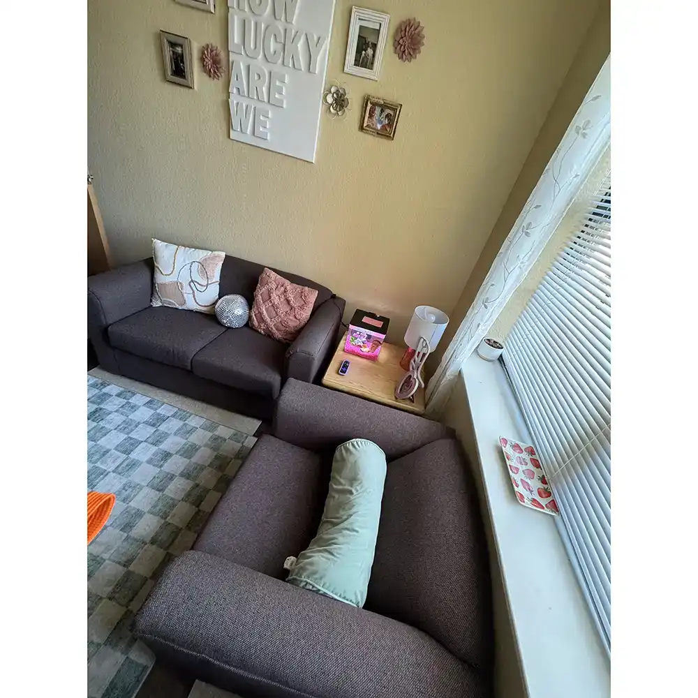 Living room in SHSU's Raven Village featuring two brown sofas arranged in an L-shape, one against the wall and the other near a window with blinds. Decorative pillows, including floral and textured designs, are on the sofa. A small wooden side table between the sofas holds a lamp, a pink box, and other items. The wall above the sofa displays framed pictures and a large sign reading 'HOW LUCKY ARE WE.' A patterned rug covers part of the floor.