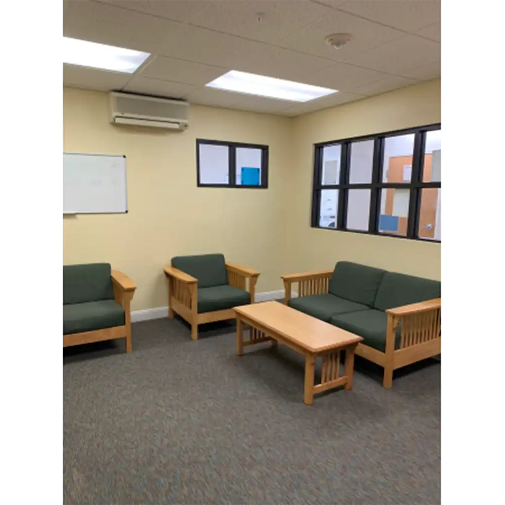 Study area in SHSU's Raven Village featuring beige walls, gray carpeted flooring, and wooden furniture with green cushions including two single chairs and a double-seater sofa. A rectangular wooden coffee table sits in front of the sofa. A whiteboard and air conditioning unit are mounted on the left wall, and two windows on the back wall look into another room or hallway. The space is lit by recessed ceiling lights.
