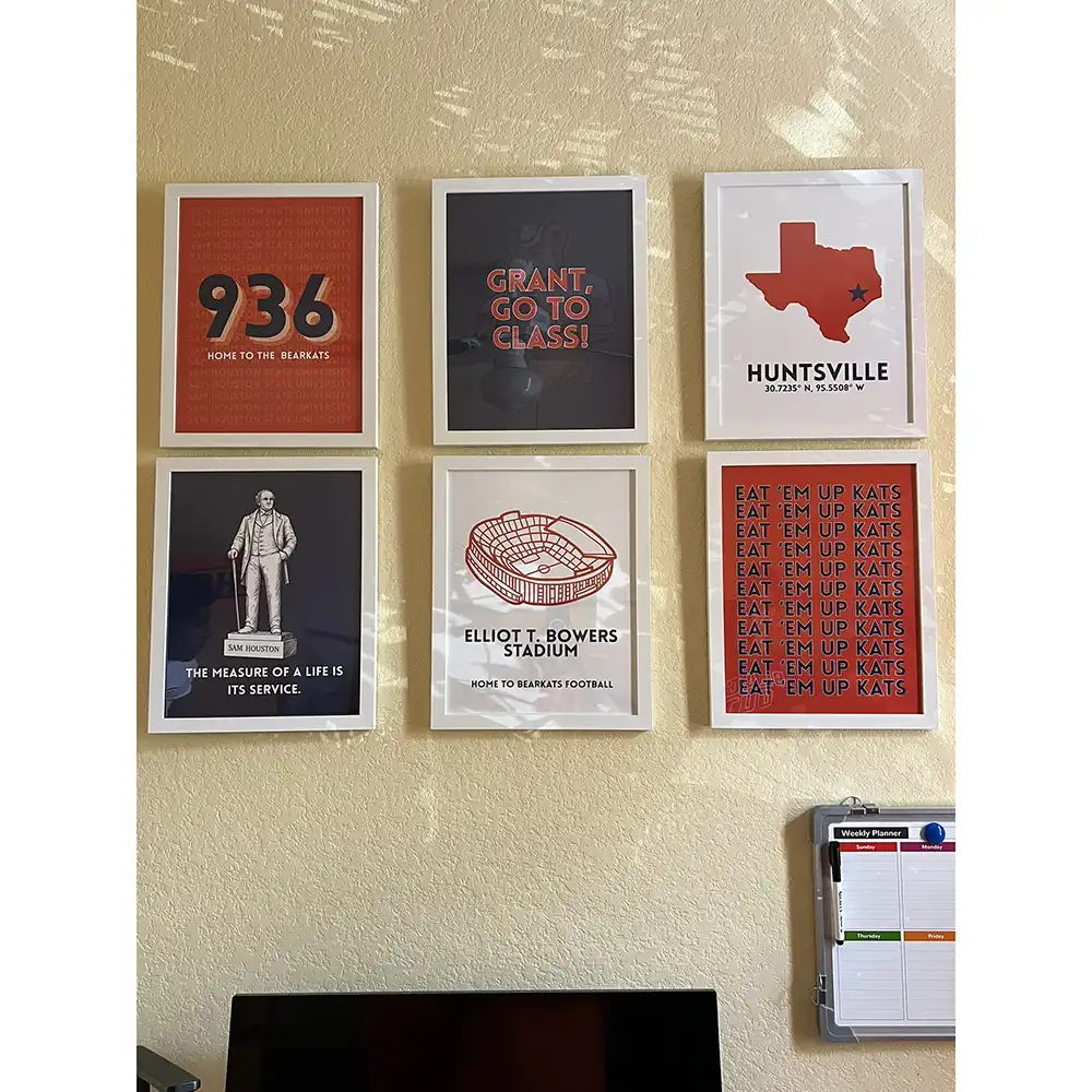 Decorated wall in a residential room at SHSU's Raven Village featuring six framed posters arranged in two rows. Top row includes: '936 Home to the Bearkats' on orange, 'Grant, Go To Class!' on dark background, and a Texas-shaped graphic with 'Huntsville' and coordinates. Bottom row includes: a Sam Houston quote poster, an image of Elliott T. Bowers Stadium labeled 'Home to Bearkat Football,' and an orange poster with the phrase 'Eat 'Em Up Kats.' A black monitor screen is partially visible below, with a weekly planner board to the right.