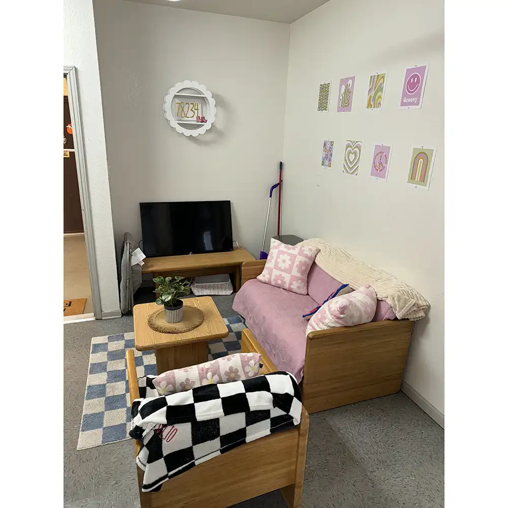 Cozy living room at SHSU's Sam Houston Village featuring a wooden sofa and loveseat with pink cushions and floral pillows, a black and white checkered blanket on the loveseat, a wooden coffee table with a potted plant, and a flat-screen TV on a wooden stand. Cleaning supplies are placed nearby, and the walls are decorated with colorful prints including rainbows, hearts, and positive messages. A blue and white checkered rug covers part of the floor.