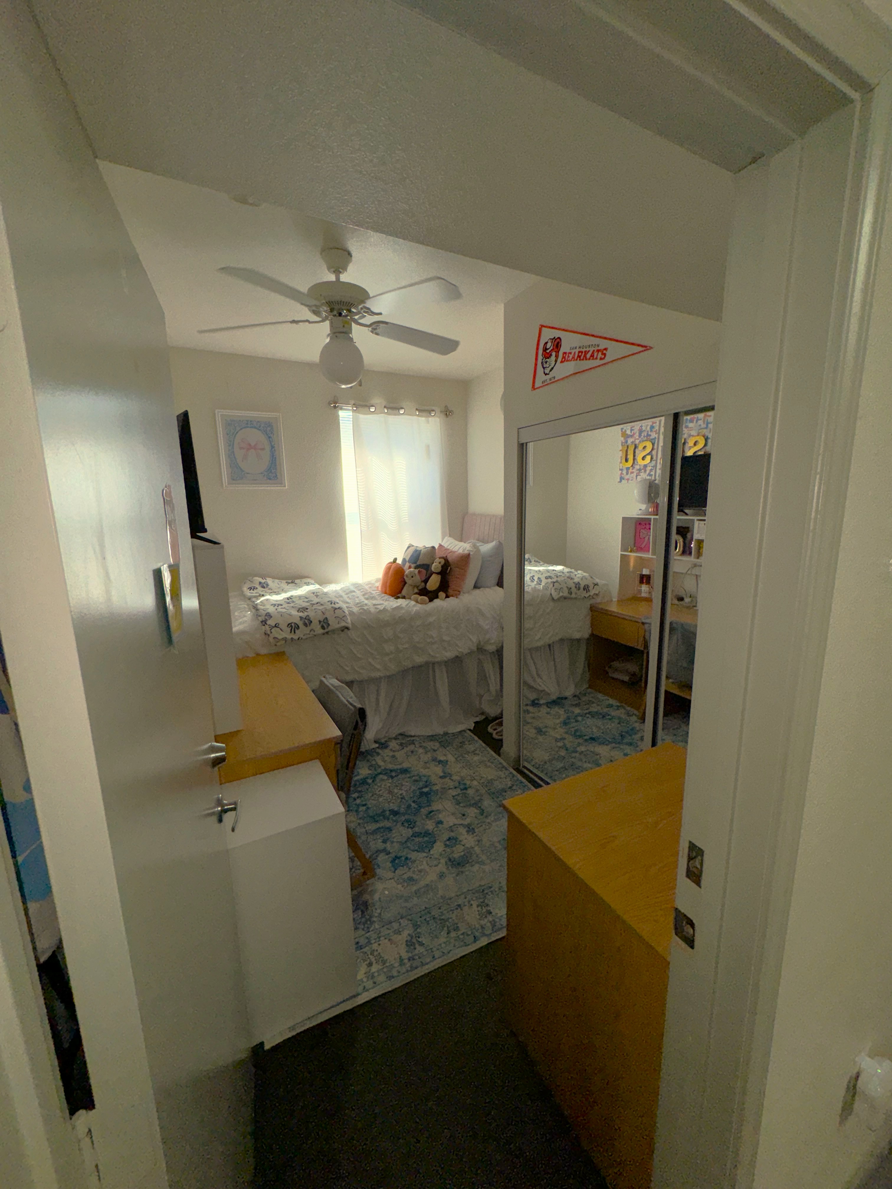 Bedroom at Sam Houston Village, SHSU, viewed from the doorway. The room features a bed with white bedding and pillows against the far wall beneath a window. A mirrored closet door is to the right, next to a desk with shelves and personal items. Another desk with a chair and storage units is on the left. The floor has a blue patterned rug, and a ceiling fan hangs above. A 'Bearkats' pennant flag is displayed near the ceiling.