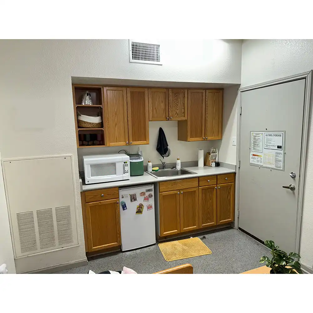 Kitchenette at Sam Houston Village, SHSU, featuring wooden cabinets above and below a countertop. A white microwave sits on a small refrigerator with magnets on the left. The center includes a stainless steel sink with cleaning supplies, and a black towel hangs above. To the right are paper towels and utensils in containers. Open shelves above the microwave hold dishes, and a nearby door displays several posted notices.