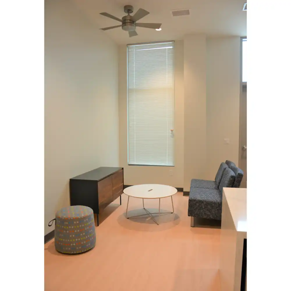 Living room of SHSU's San Jacinto Studio Apartments with modern decor, light-colored walls and flooring. Furnishings include a ceiling fan, large window with blinds, blue patterned ottoman, wooden cabinet with black accents, round white coffee table, and a gray upholstered sofa.