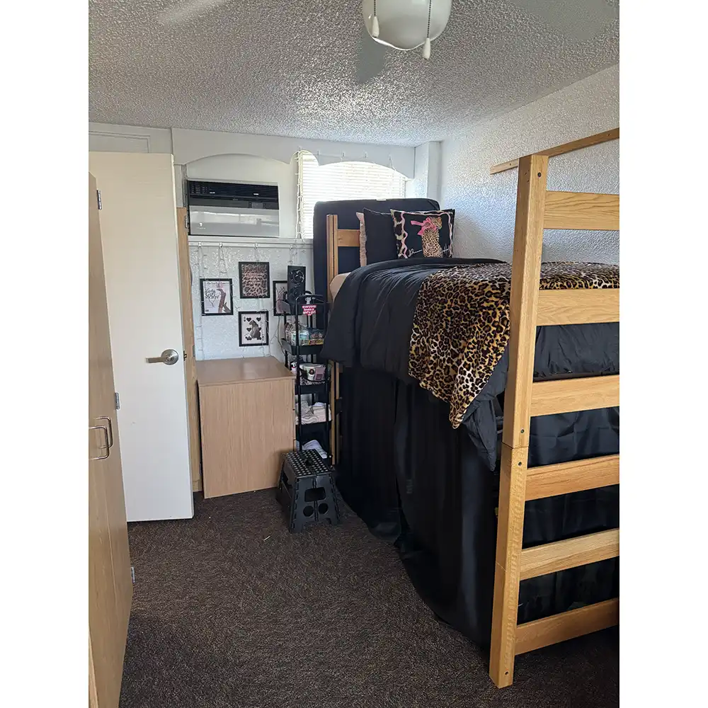Bedroom in SHSU’s White Hall with a lofted bed featuring black and leopard print bedding. Another bed sits below, alongside a small shelving unit. The room has brown carpet, white walls, framed wall art, a window with an air conditioning unit, and a ceiling fan. Not pictured is a 3rd twin XL bed. White Hall is currently set up to sleep 3 to a bedroom.