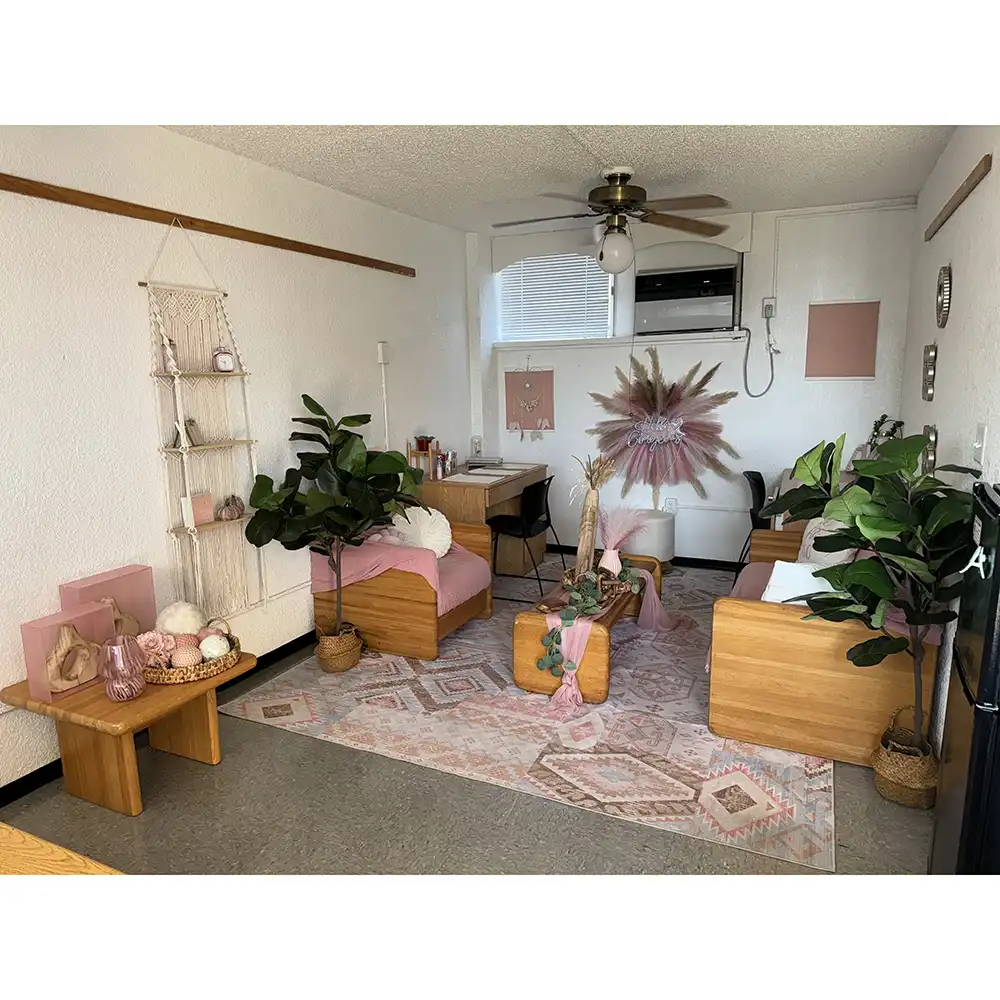 Living room in SHSU’s White Hall featuring wooden furniture with pink cushions, a pink and white patterned rug, potted plants, a desk with decorative items, and wall art including a macramé hanging. A ceiling fan with a light and a window-mounted air conditioner are also visible.