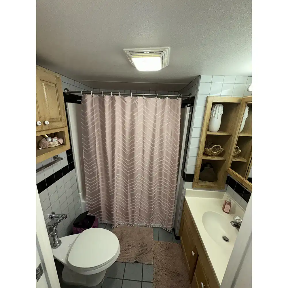 Bathroom in SHSU’s White Hall with a white toilet, pink chevron shower curtain, gray tile flooring, and multiple pink bath mats. A wooden vanity with a white sink and open shelving holds toiletries and towels. White tiled walls with black accents and an overhead light complete the space.