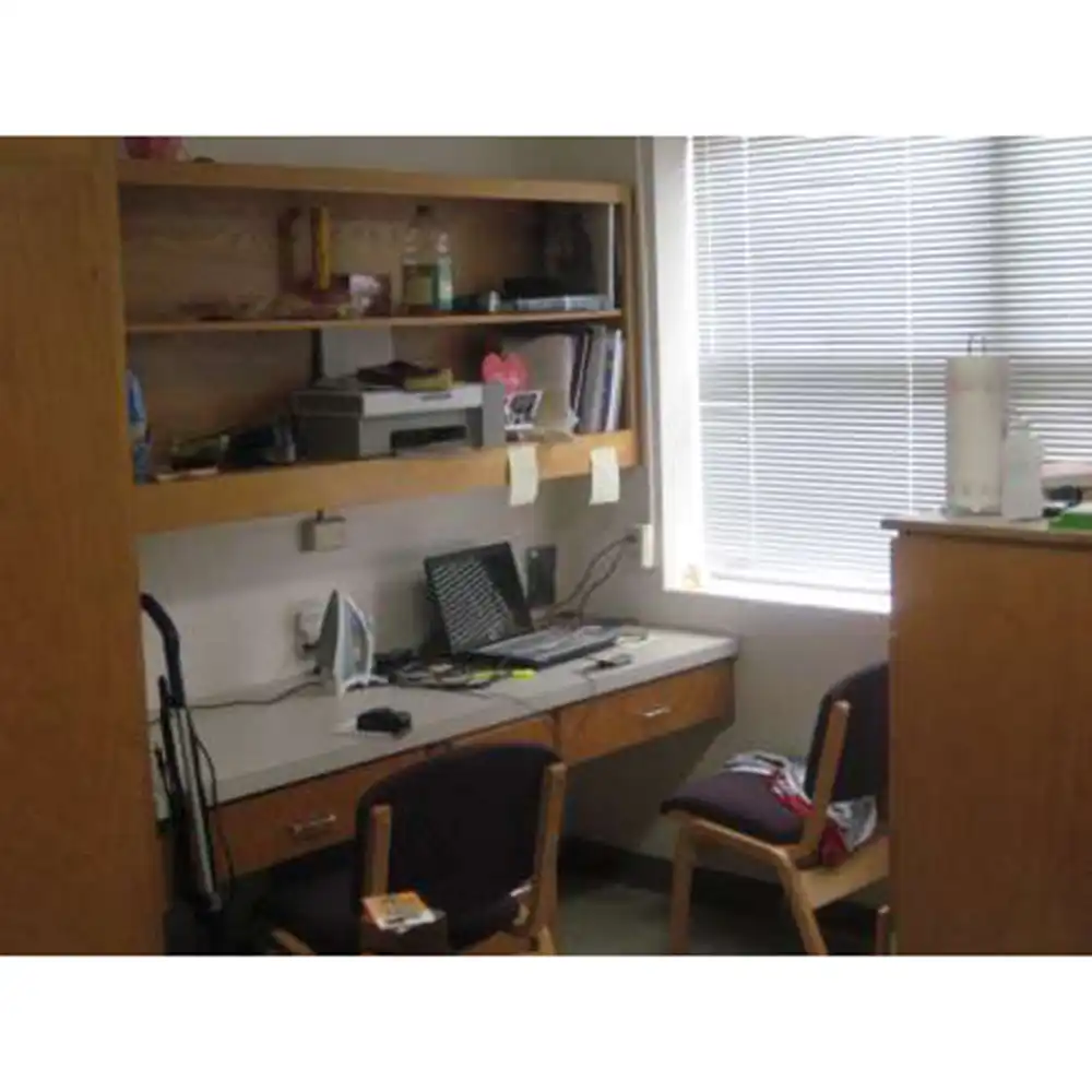 Bedroom in SHSU's Small Houses featuring a built-in wooden desk with drawers and an overhead shelf holding books, a water bottle, and personal items. The desk surface includes an iron, a laptop, electronic devices, and sticky notes. Two chairs sit in front of the desk, one with clothing draped over it. A window with closed blinds is to the right, allowing natural light into the room.