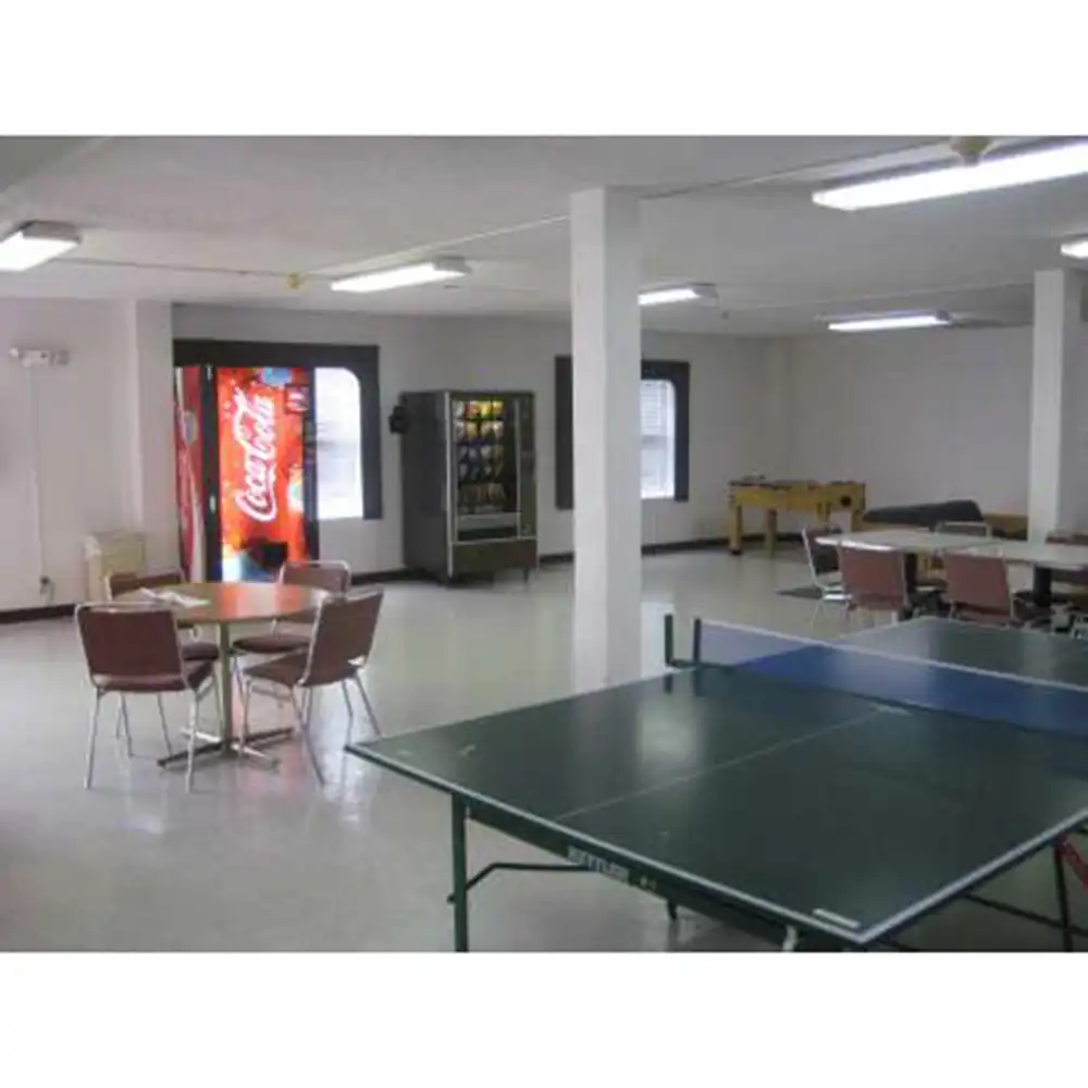 Lobby in SHSU's Small Houses featuring a green ping pong table in the foreground, two round tables with chairs to the left, and a Coca-Cola vending machine beside a snack machine against the far wall. The room also includes a foosball table and additional seating areas, with white walls, tiled flooring, and fluorescent lighting.