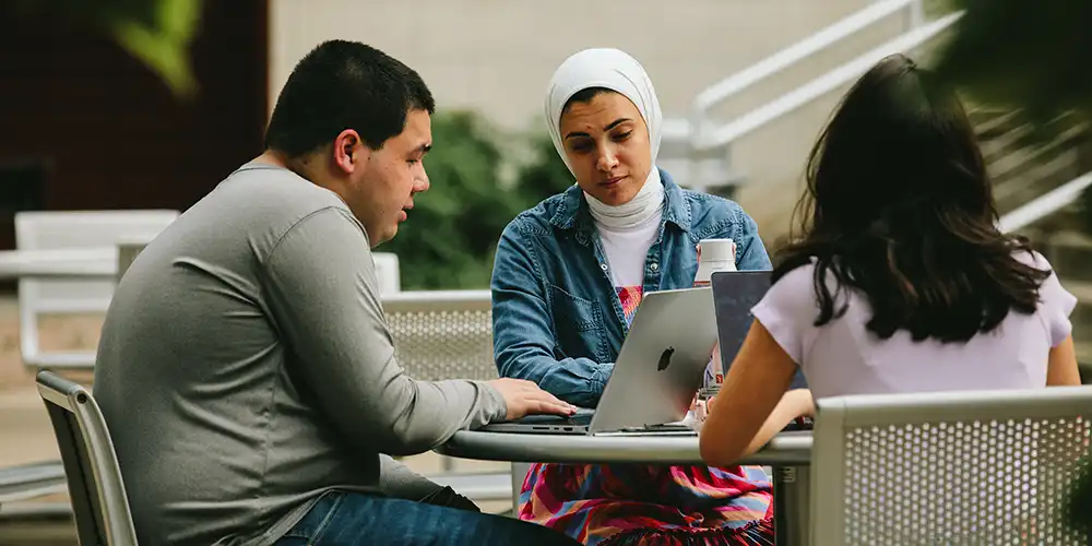 Three students sitting at an outdoor table, working together on laptops and notes, engaged in discussion.