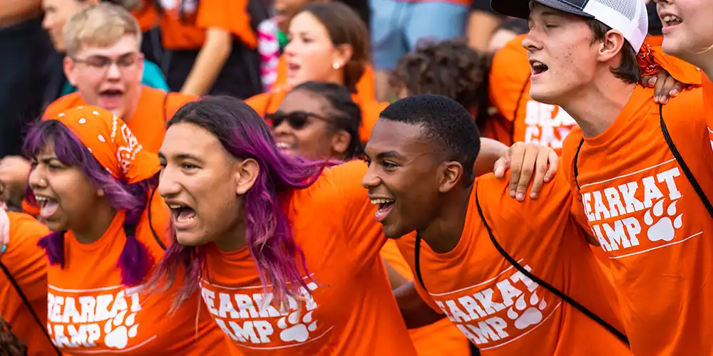 A large group of enthusiastic young people, all wearing bright orange "BEARKAT CAMP" T-shirts, participating in a college spirit event. Many are arm-in-arm, crouched down in the front chanting, while others in the background cheer and gesture excitedly.