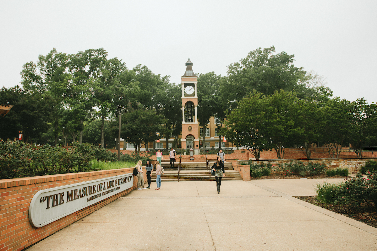 Frank Parker Yard with the SHSU motto on a curved brick wall and the clock tower in the background as students gather.