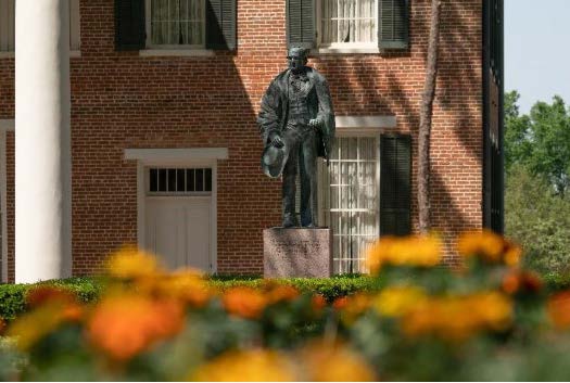 Bronze statue of Sam Houston outside Austin Hall, surrounded by a garden with bright orange and yellow flowers.