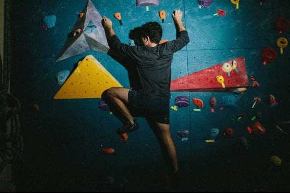 A rock climber pauses on a blue climbing wall, studying the route among various colored holds to decide his next agile move.