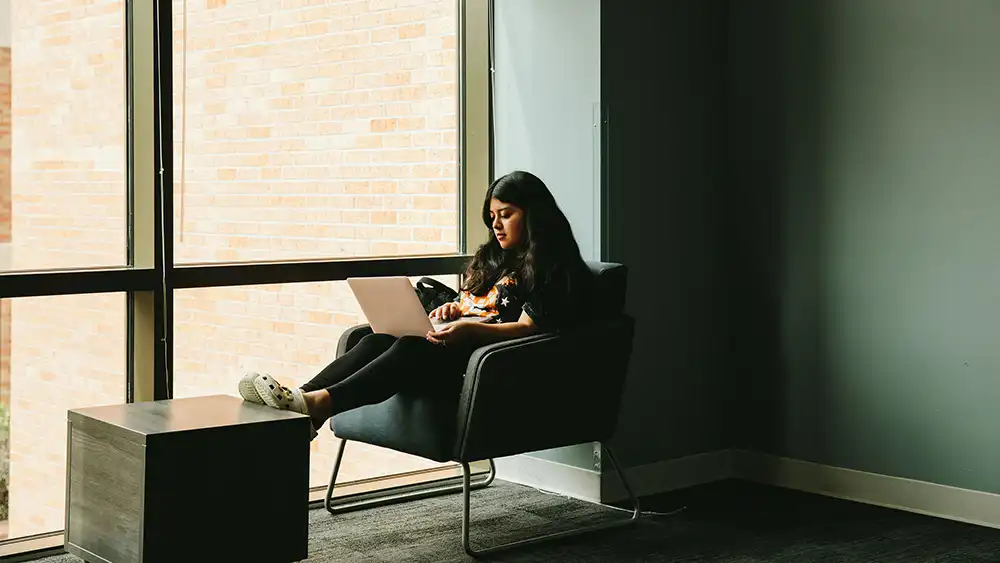 An image depicting a student sitting in a modern lounge area. The student is seated in a dark, cushioned chair by a large window, letting in natural light. The student, wearing a black T-shirt with orange and white accents and white Crocs, is using a laptop. A small, dark coffee table sits in front of the student. The lounge area features a muted color palette, including dark carpet, gray walls, and contemporary furniture. The overall atmosphere is calm and relaxed.